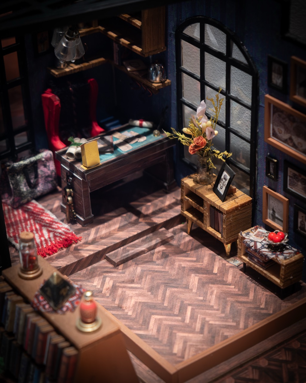 A cosy, miniature room with a chevron wood floor, a desk by a large arched window, bookshelves, framed pictures on deep blue walls, a red rug, and small furniture and decor details throughout.