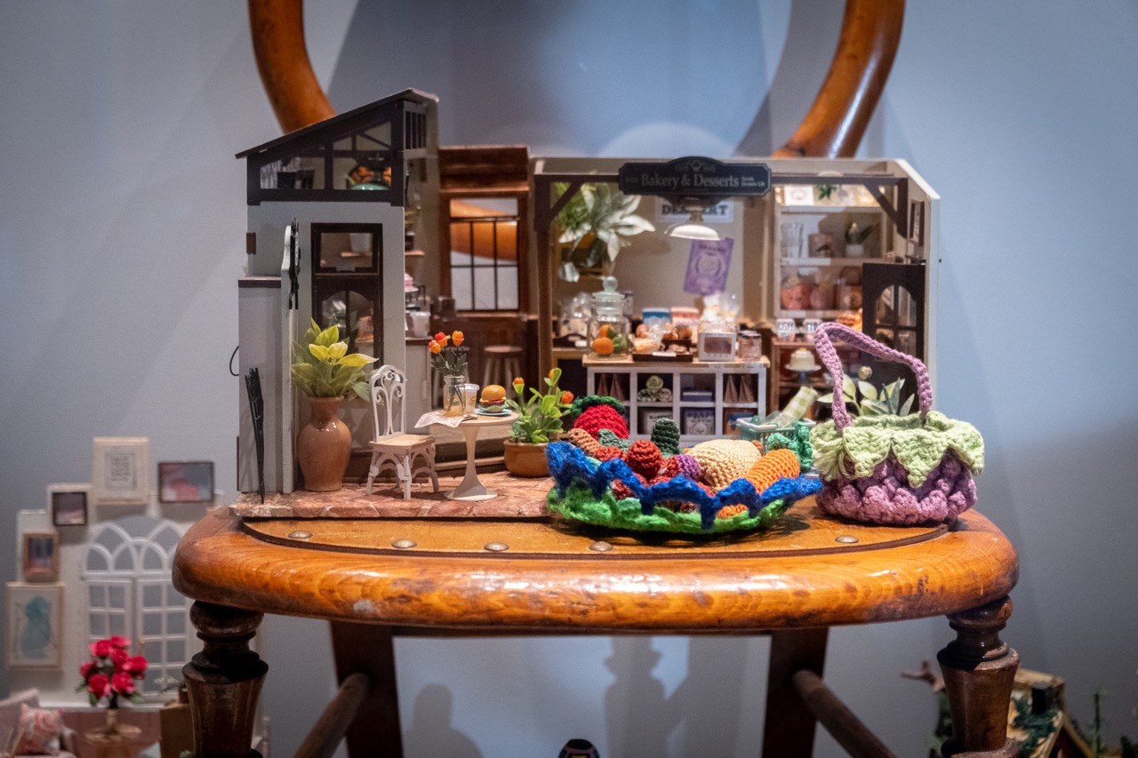 A miniature bakery diorama with tiny furniture and detailed pastries is displayed on a wooden chair. Colourful crocheted decorations resembling fruit sit in the foreground. The background is softly blurred.