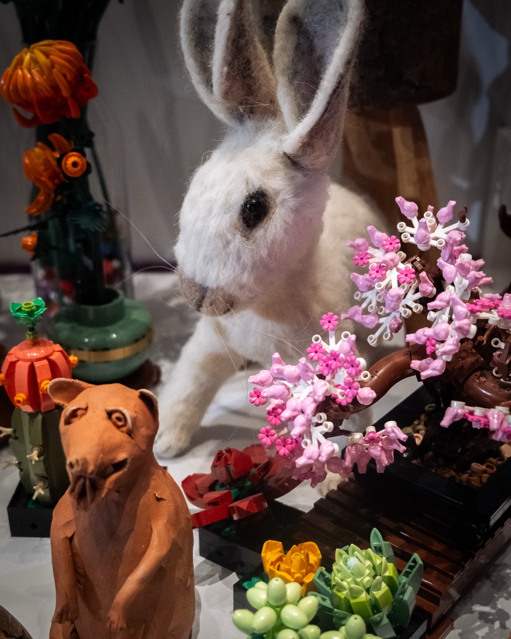 A felted white rabbit with upright ears is surrounded by colourful clay and LEGO flowers, succulents, and a small brown clay animal figure on display.