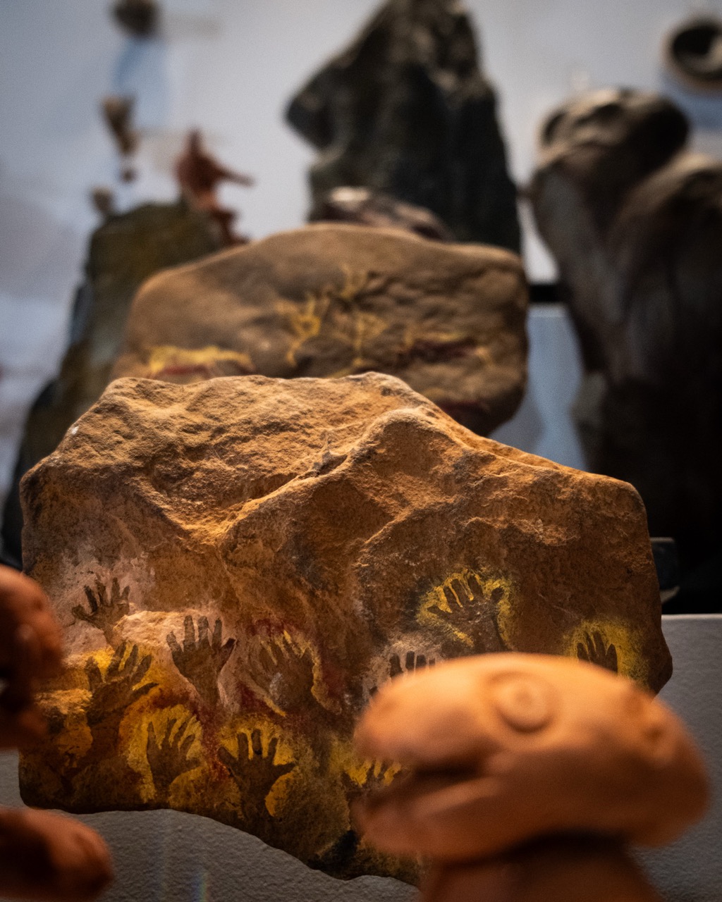 A large rock with ancient handprint art in yellow pigment is displayed in a museum, surrounded by other rocks and artefacts in the background.