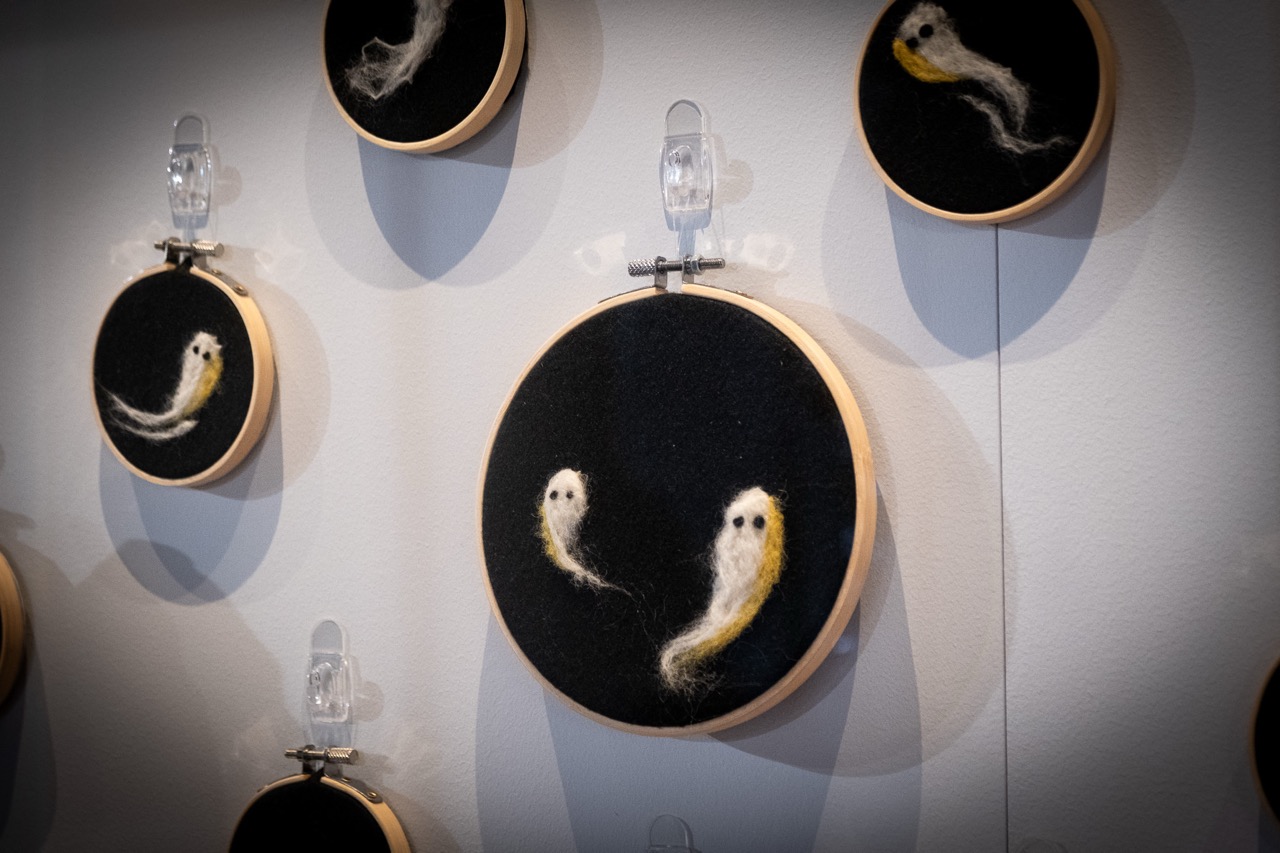 Several embroidery hoops hang on a wall, each displaying felted art of small, ghost-like figures on a black background. The hoops are attached with clear plastic clips.