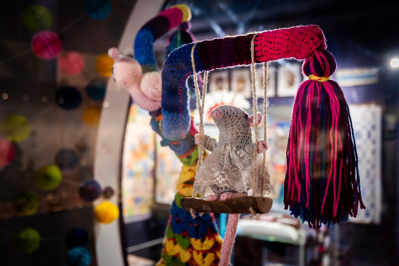 A small, hand-knitted mouse wearing a lace dress sits on a swing made of yarn and wood. The swing is suspended from a colourful, crocheted structure. The background is filled with colourful decorations and blurred lights.