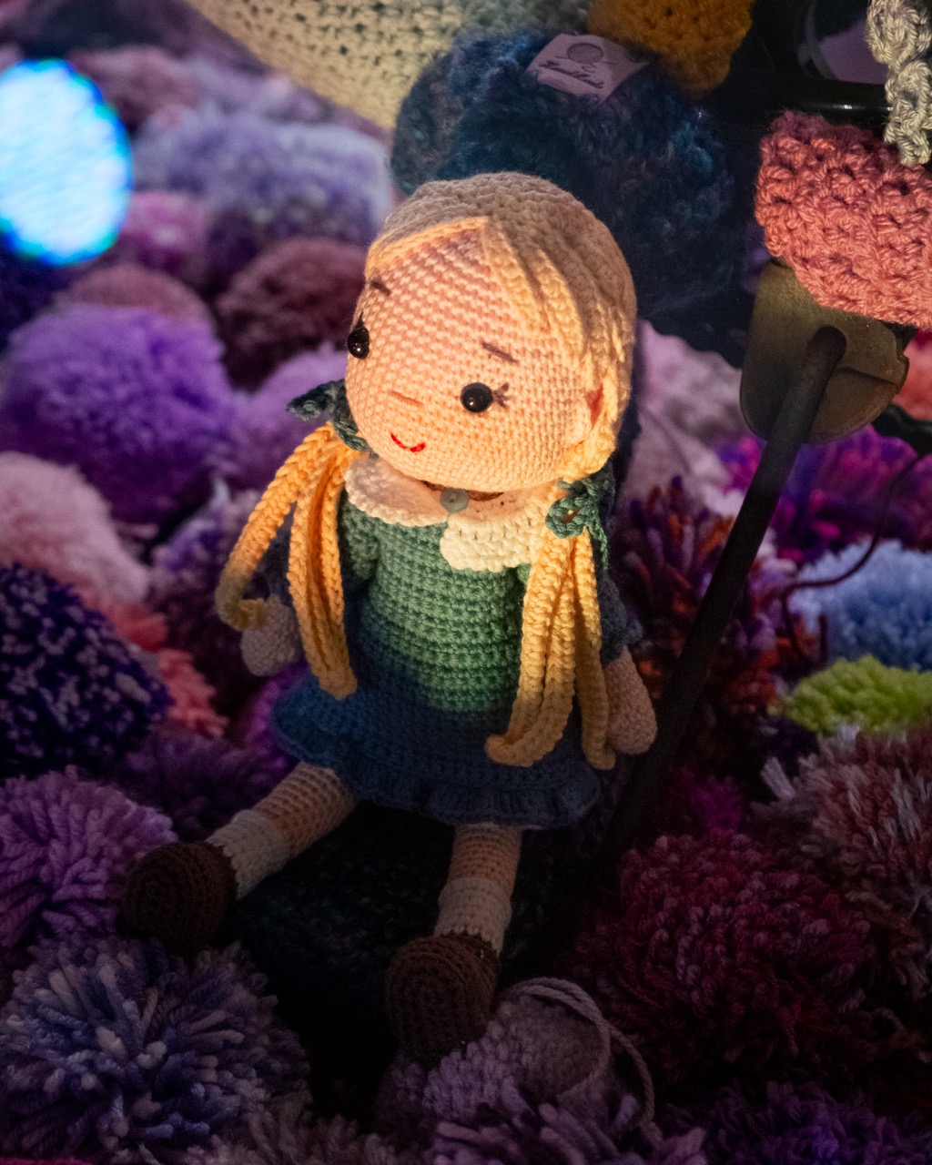 A crocheted doll with long blonde hair, wearing a blue dress and a white collar, sits surrounded by colourful, fluffy yarn pom-poms in purples, pinks, and blues, illuminated by soft lighting.