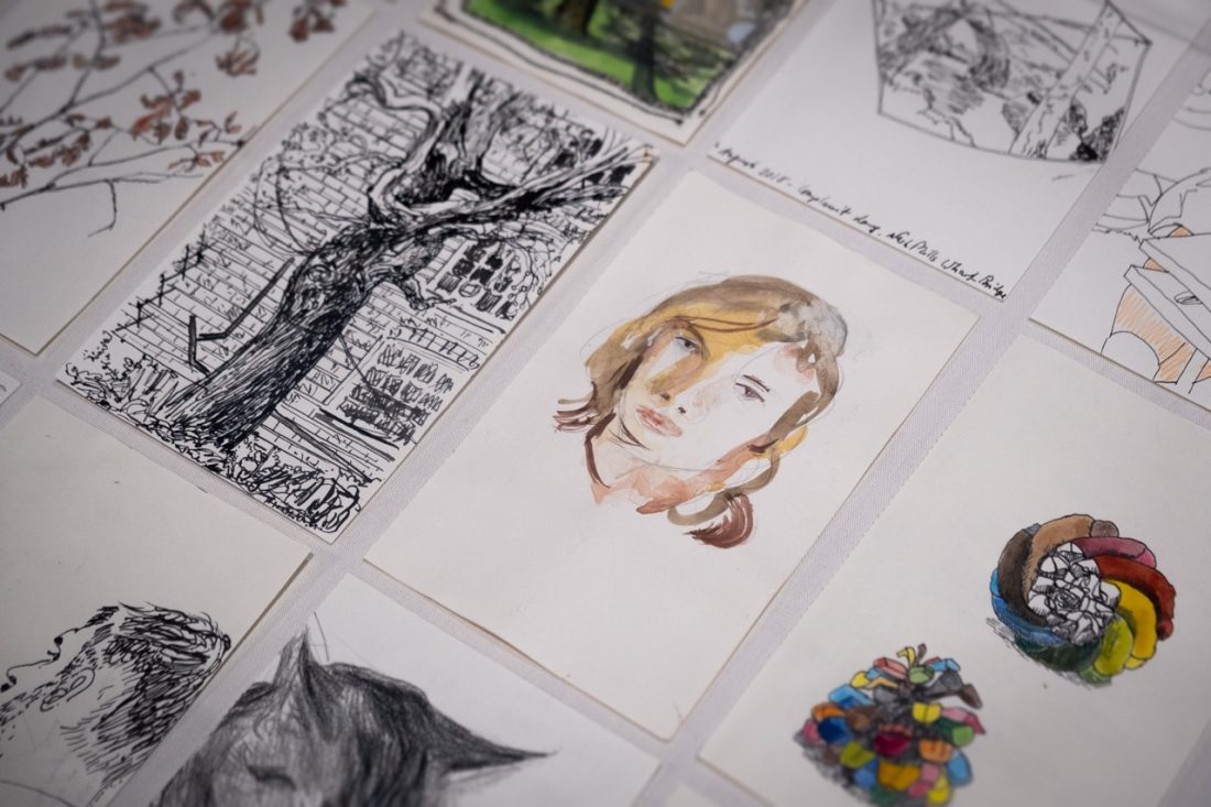 A collection of hand-drawn and painted art pieces is laid out, featuring sketches of trees, faces, and colourful objects on white paper. The central focus is a watercolour portrait of a person's face.