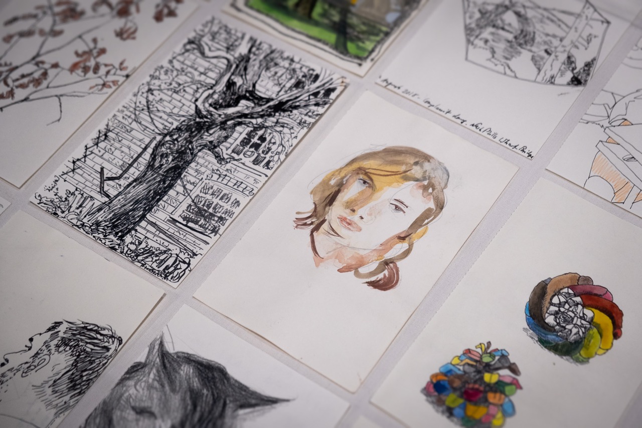 A collection of hand-drawn and painted art pieces is laid out, featuring sketches of trees, faces, and colourful objects on white paper. The central focus is a watercolour portrait of a person's face.