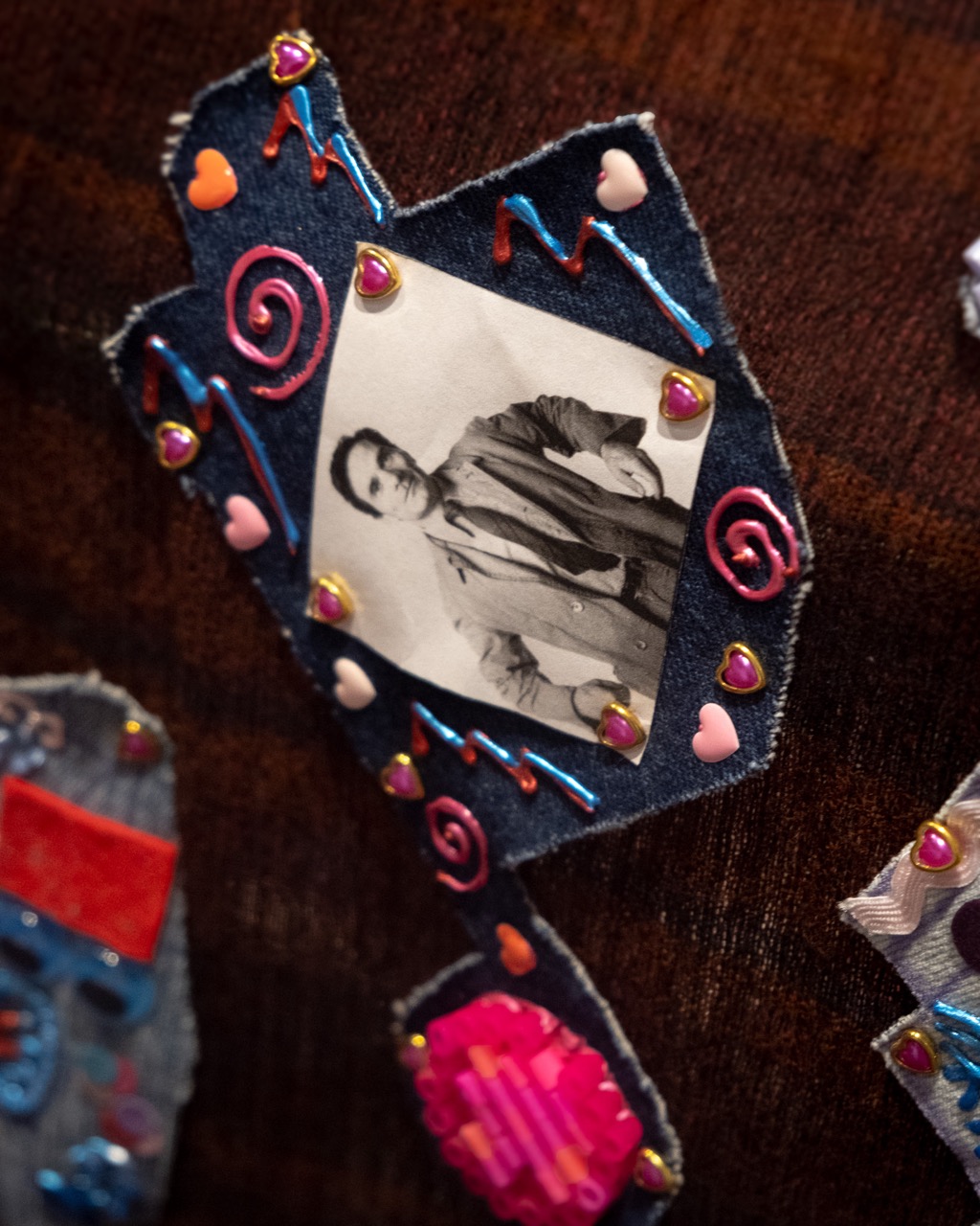 A black-and-white photo of a man is attached to a denim dreidel shape, decorated with beads, heart-shaped sequins, and pink and blue embellishments on a dark fabric background.