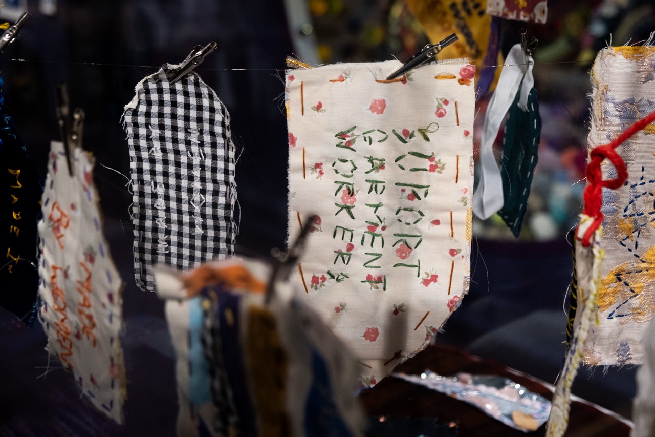 A close-up of small fabric squares hanging on a string with pegs. One piece has embroidered text, another has a black and white chequered pattern, and others have colourful designs.