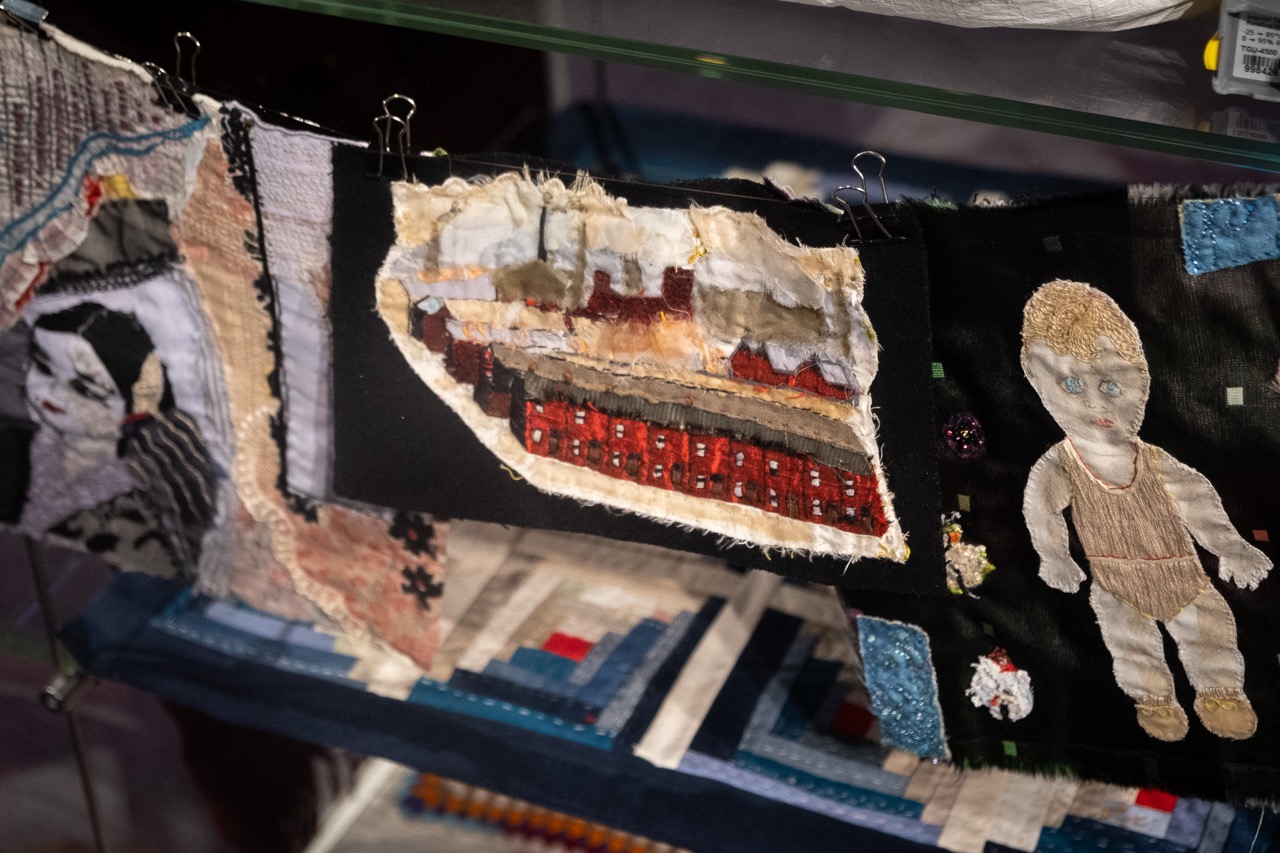 Textile art panels clipped to a display shelf, featuring fabric collages: a stitched female face, a cityscape with red buildings on black fabric, and an embroidered child figure on black cloth.