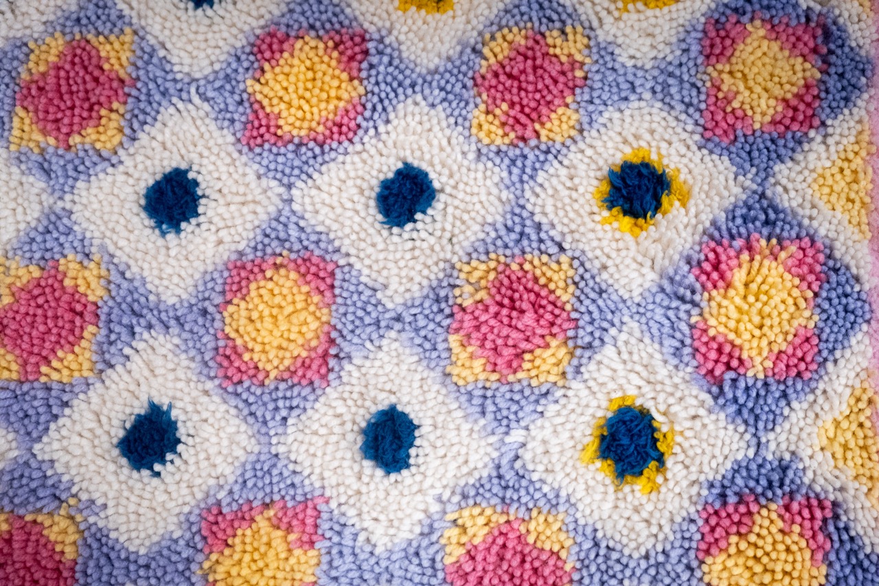 A close-up of a textured, multicoloured crochet or tufted fabric featuring a repeating pattern of squares with blue, pink, and yellow centres surrounded by white and light purple detailing.