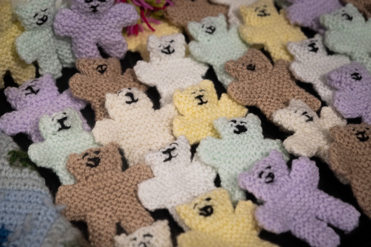 A collection of small, hand-knitted teddy bears in various pastel colours, including yellow, white, beige, purple, and mint green, arranged closely together.