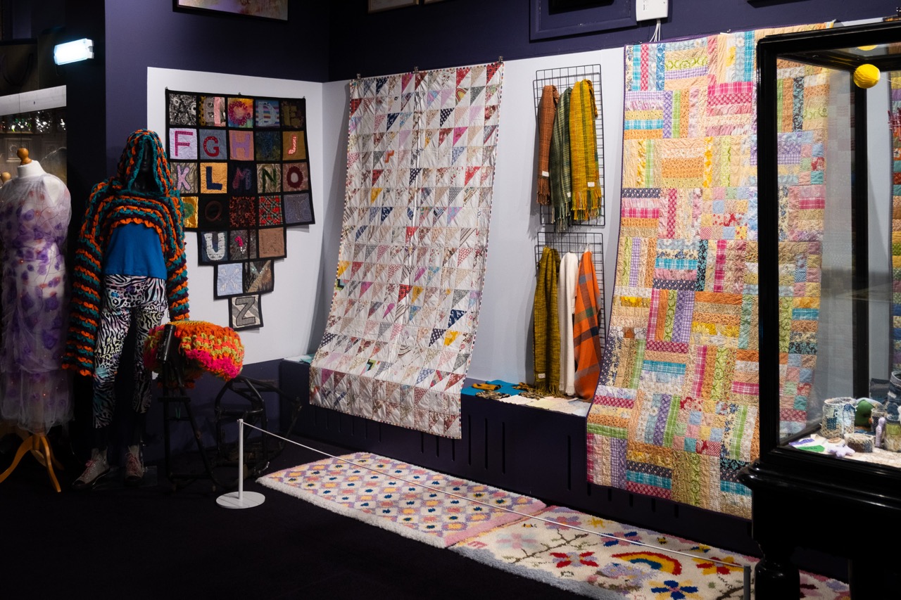 A museum display features colourful quilts hanging on the wall, a mannequin draped in a vibrant crocheted shawl, framed textile art, and various knitted scarves, with a patterned rug and floral stool in front.