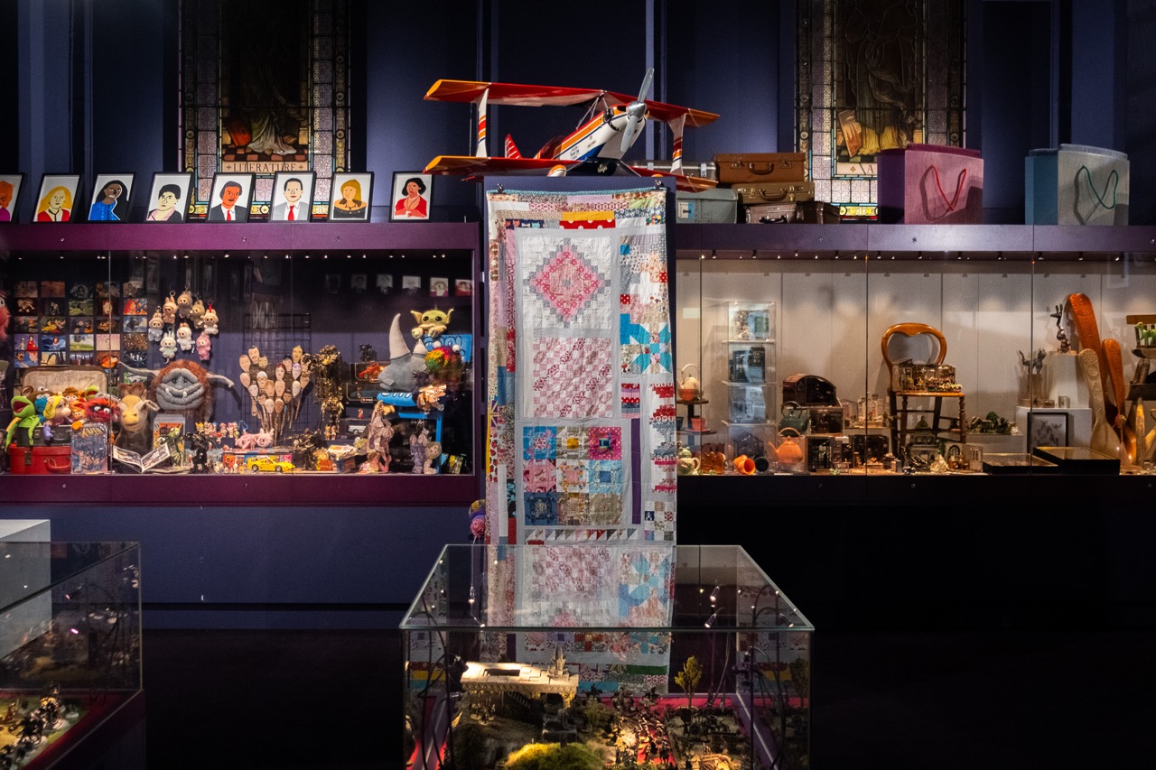 A museum display features colourful quilts, model aeroplanes, dolls, and various toys and trinkets in glass cases. The background includes framed portraits, souvenirs, and decorative objects on shelves.
