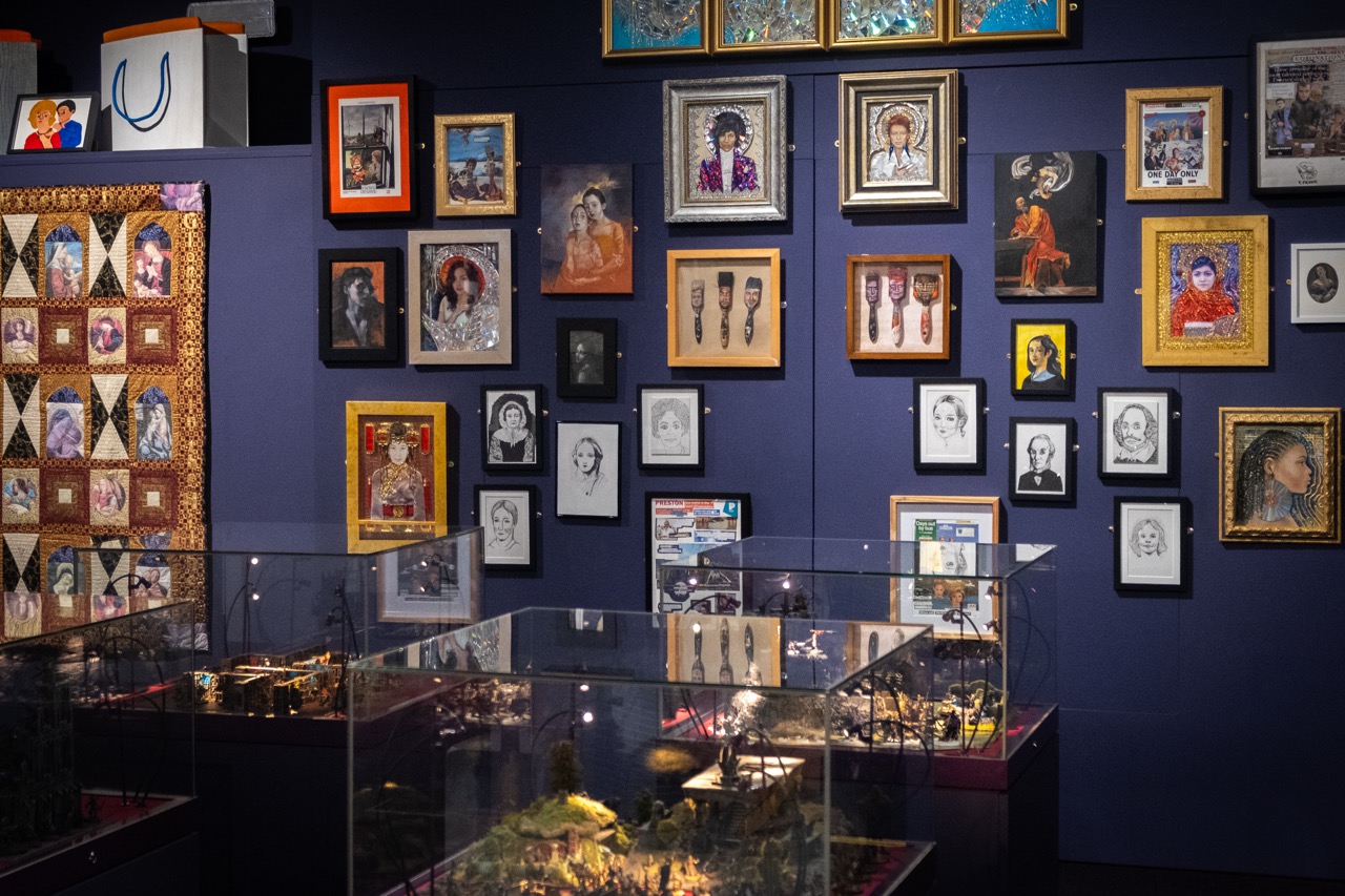 Gallery wall with numerous framed artworks and portraits of diverse people. Glass cases below display miniature models or dioramas. The wall and decor are colourful, with a quilted textile piece visible on the left.