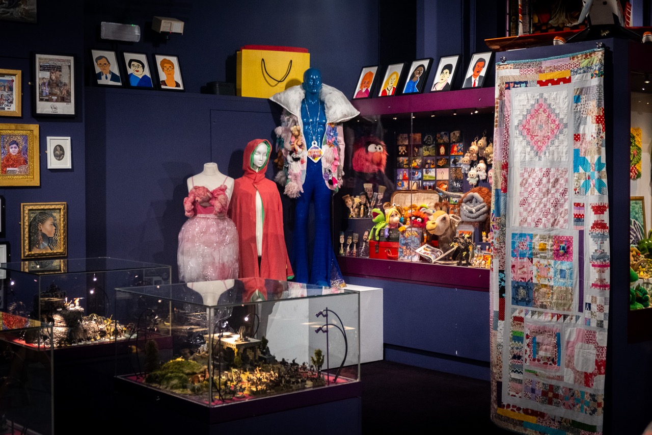 A colourful museum display with mannequins in costumes, toy collections in glass cases, framed art on deep blue walls, and a patchwork quilt hanging on the right side.