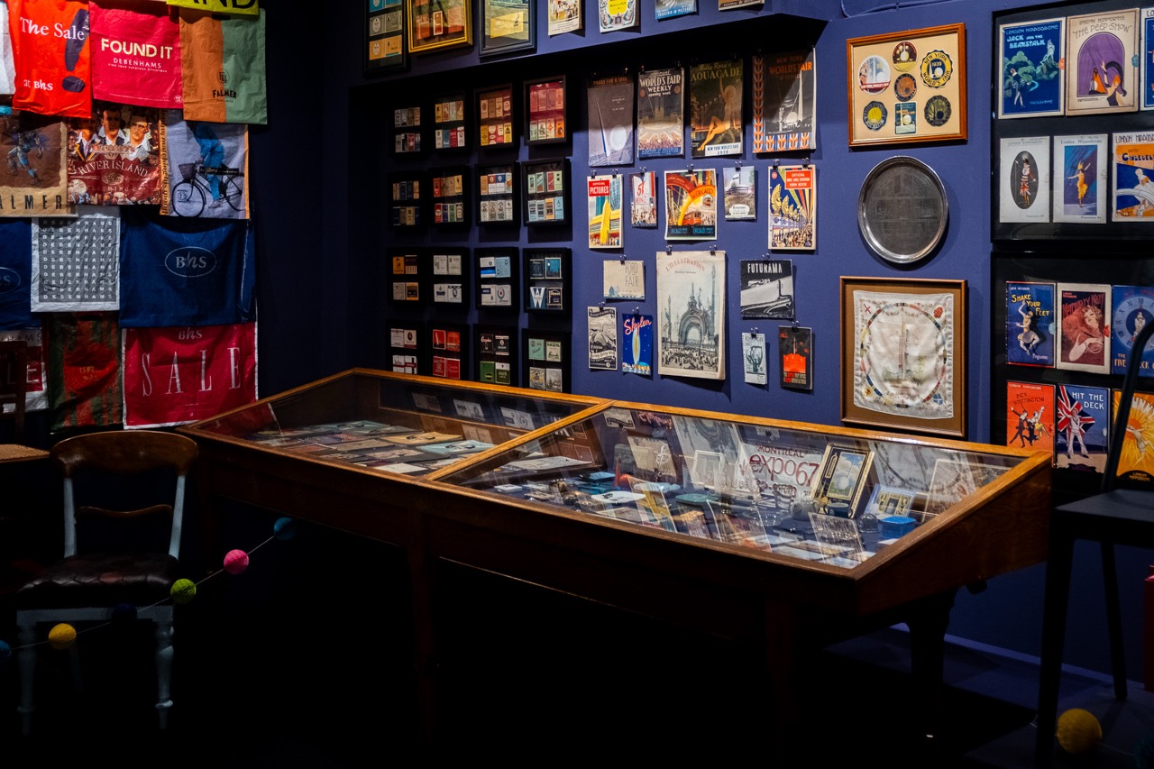 A display room with glass cases filled with memorabilia and collectables, walls covered with vintage posters, postcards, and colourful bags, creating a museum-like atmosphere.