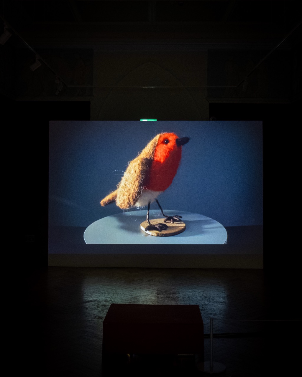 A projection shows a close-up image of a wool felt bird with a red breast and brown body, standing on a circular base against a blue background in a dark exhibition space.