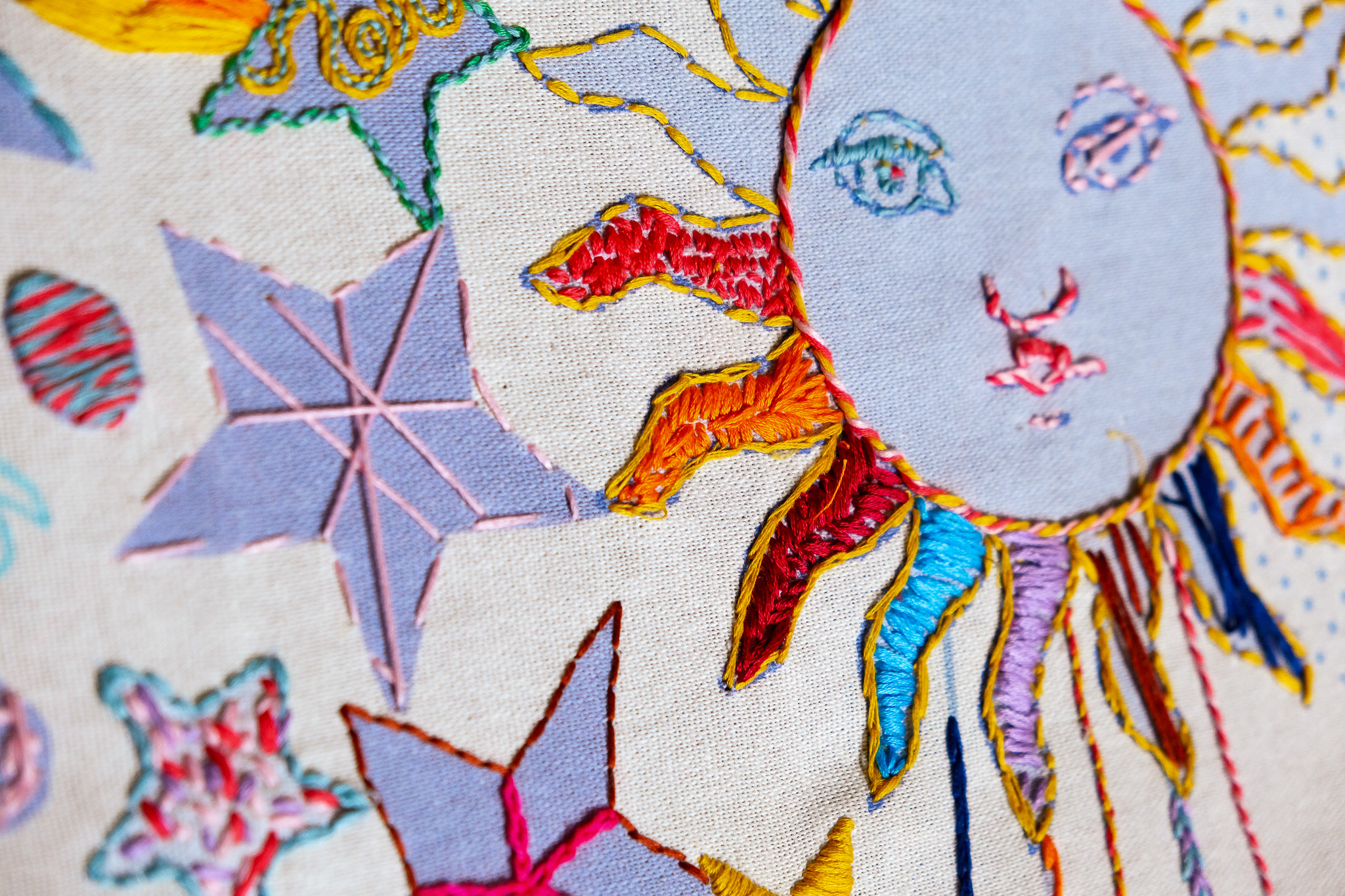 Colourful embroidery features a sun with a face, using vibrant threads on fabric.