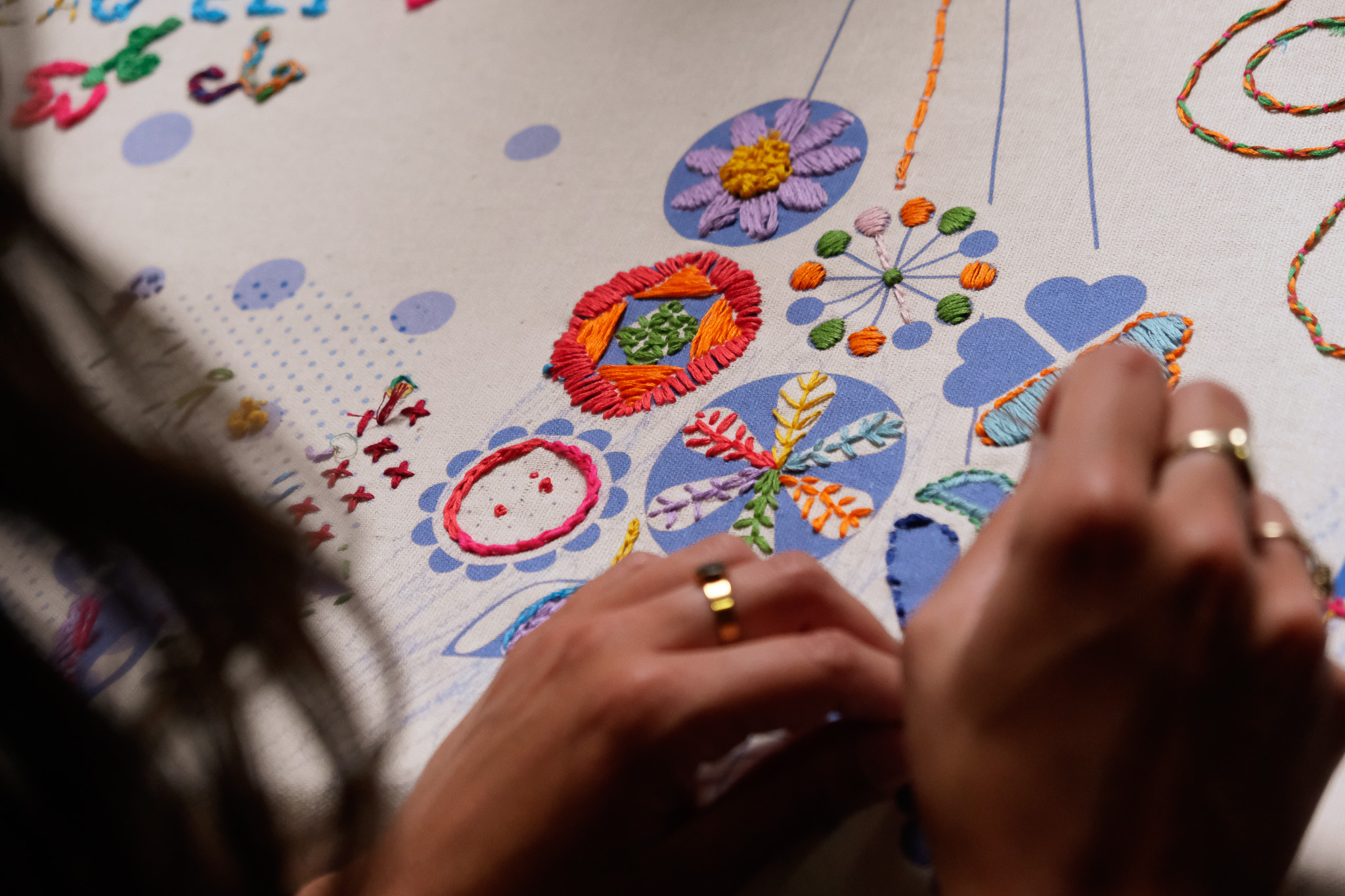 Close-up of hands with rings embroidering colourful floral patterns on a white fabric, using various embroidery stitches and threads to create detailed, vibrant designs.