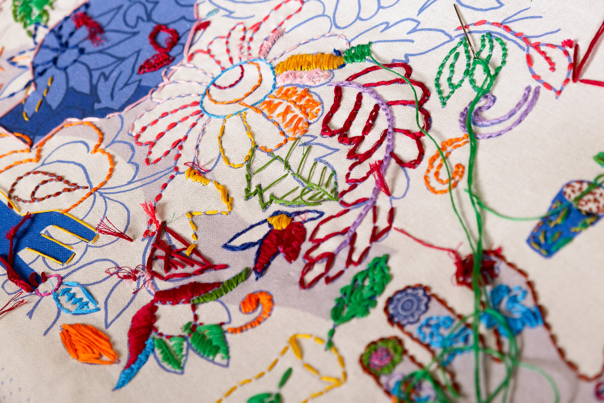 Close-up of colourful embroidery on fabric featuring vibrant flowers and leaves. Stitches in red, green, orange, and blue.
