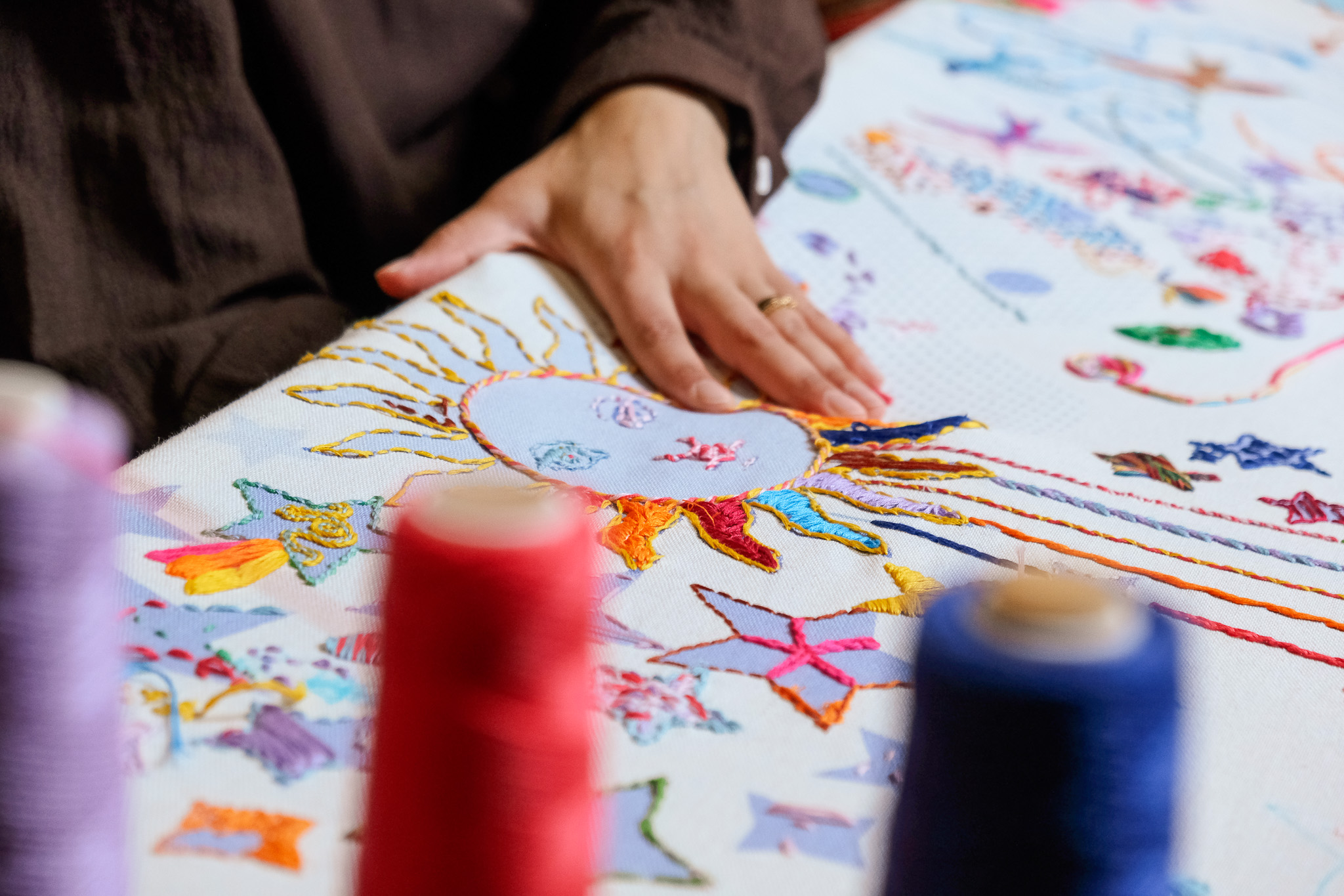 A person’s hand rests on a vibrant embroidered fabric featuring a sun and various colourful designs, with spools of thread in the foreground.