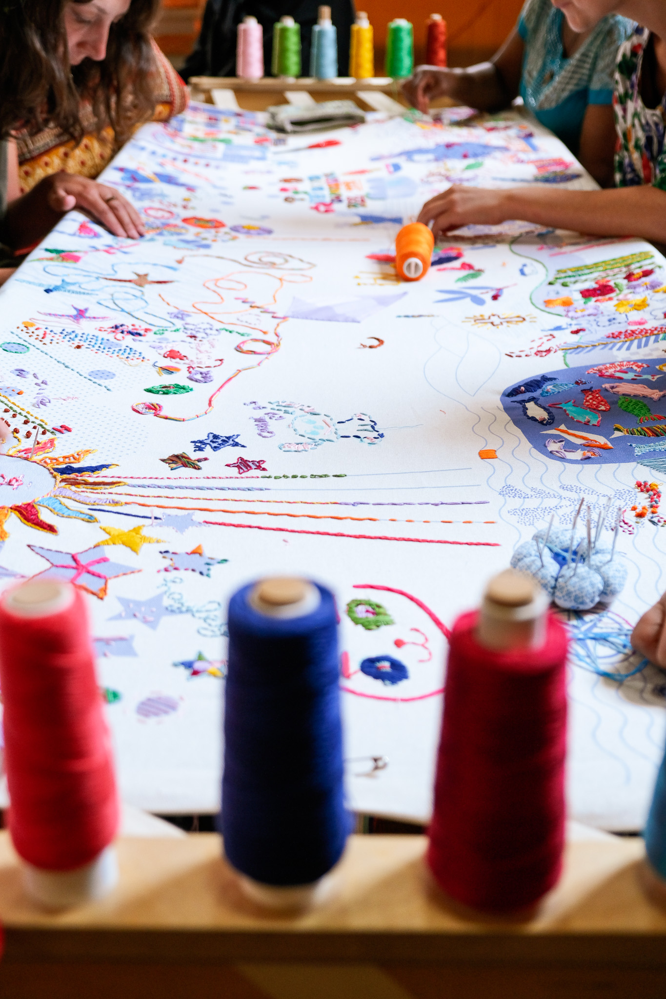 Several people sit round a table working on a large, colourful embroidery project. Reels of thread in various colours are in the foreground, with intricate stitched patterns visible on the fabric.