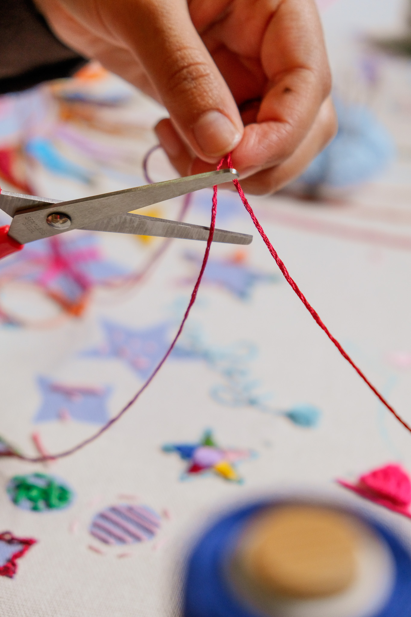 A close-up of a hand holding scissors, cutting a piece of red string. Colourful decorations and craft materials are scattered on a white surface in the background.