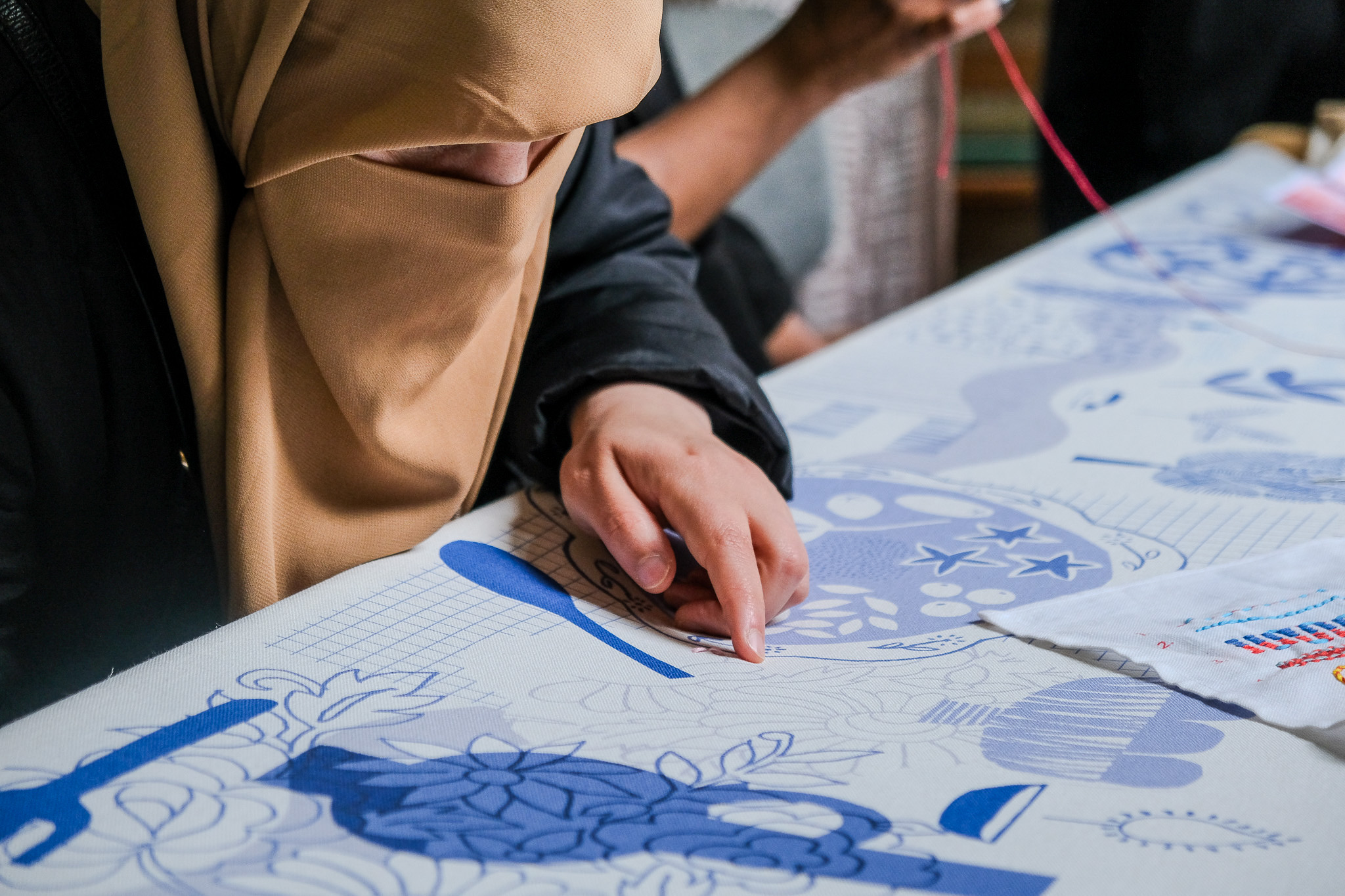 A person wearing a tan hijab and black jacket is focused on detailed embroidery work, stitching patterns on white fabric with blue designs.