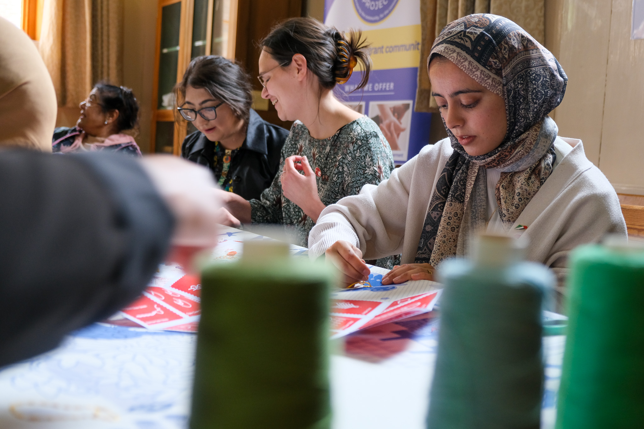 Four women sit at a table working on crafts with red paper. Reels of green thread are in the foreground, and the women appear focused and engaged in their activity.