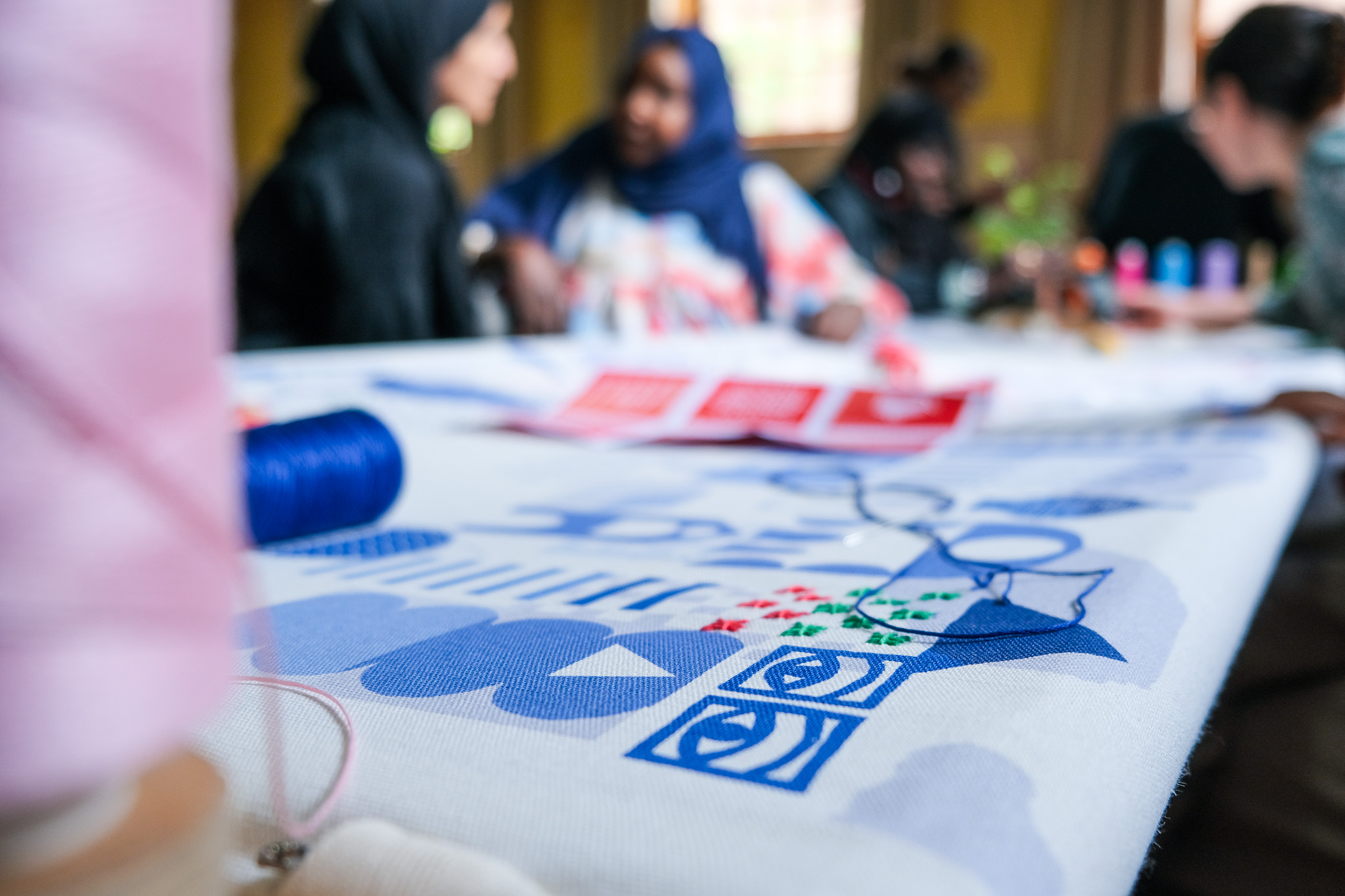 A close-up of a colourful embroidery project on a table, with blurred people in the background engaging in conversation. Spools of blue and pink thread are visible, along with patterned fabric and printed designs.