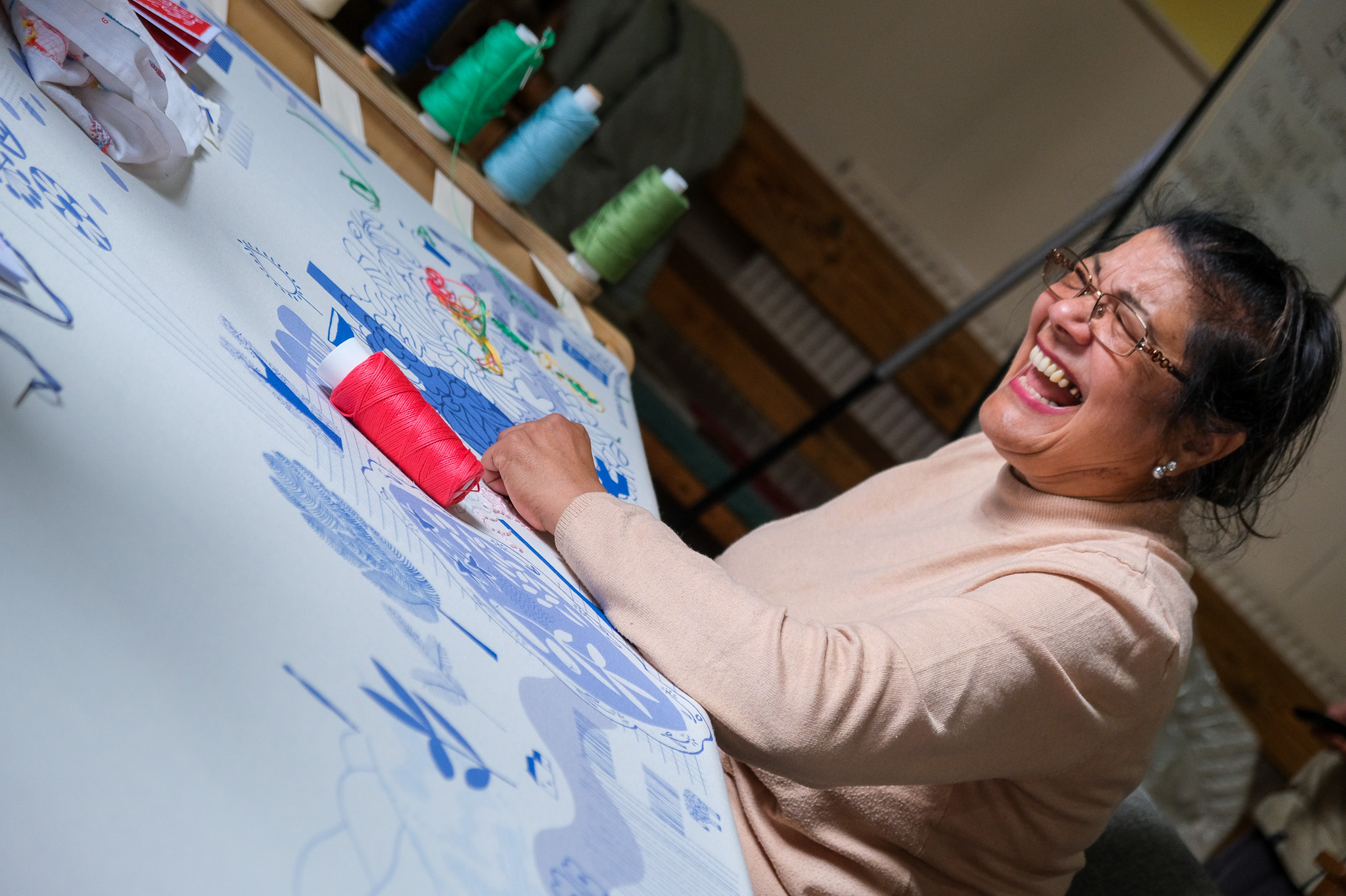A woman wearing glasses and a beige jumper sits at a table with colourful spools of thread, laughing joyfully whilst working on a fabric art project.