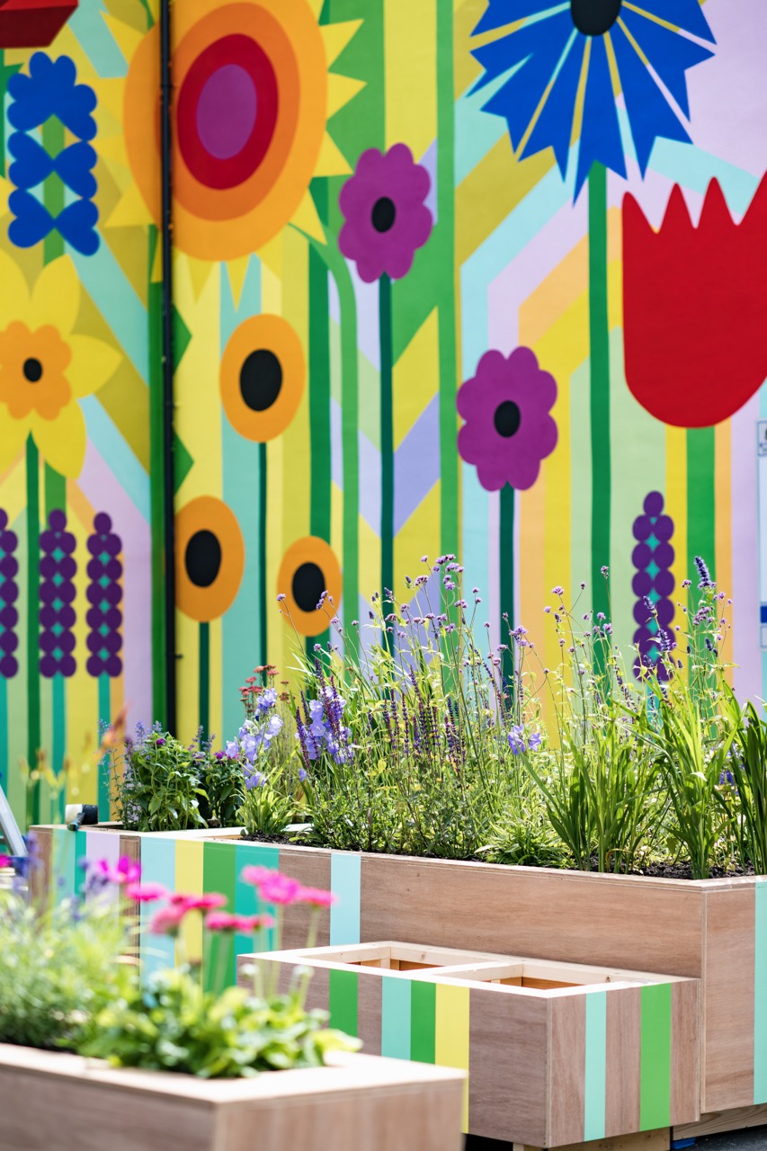 A colourful mural of oversized flowers covers a wall behind wooden planter boxes filled with blooming flowers and green plants. The bright mural and vibrant plants create a cheerful, lively outdoor scene.