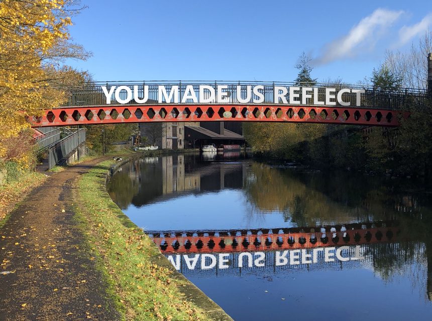 A red iron bridge over a calm canal displays large white letters reading YOU MADE US REFLECT, with the words and autumn trees reflected in the water below.