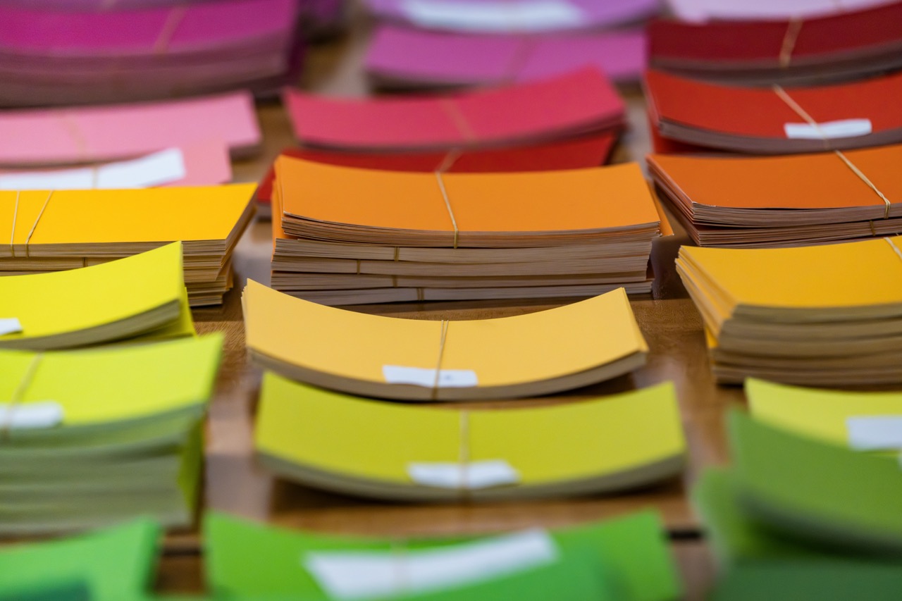 Stacks of colourful paint swatches in shades of yellow, orange, red, pink, and green are arranged in rows on a wooden surface, with each stack bound together by an elastic band.