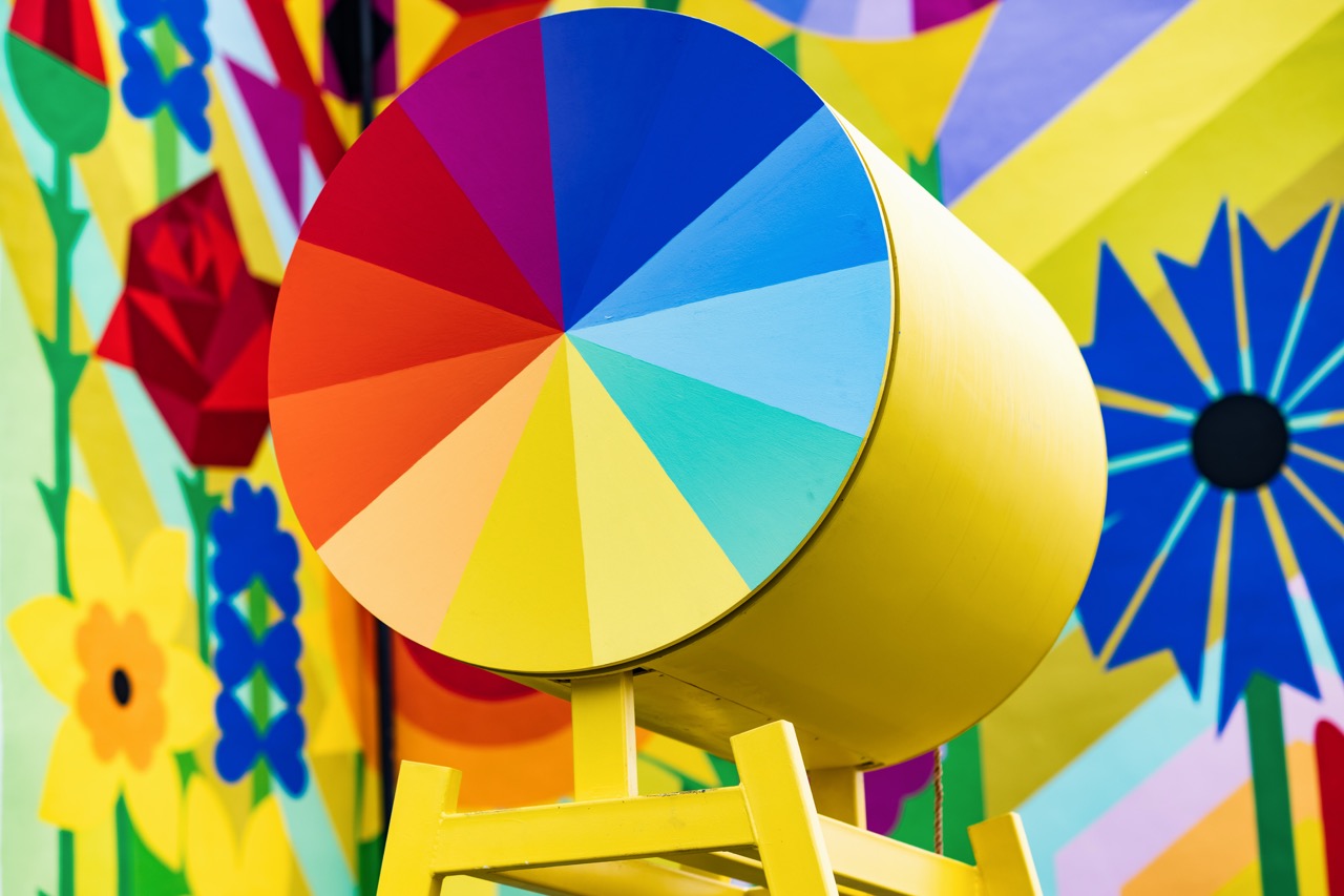 A large yellow cylinder with a circular front divided into colourful rainbow segments stands in front of a vibrant mural with bold flowers and geometric shapes in red, blue, yellow, and green.