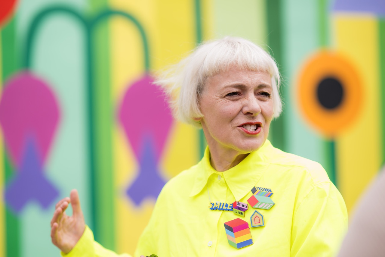 A person with short, white hair wears a bright yellow shirt and colourful pins, smiling and gesturing with one hand. The background features a vibrant, abstract mural with bold shapes and colours.