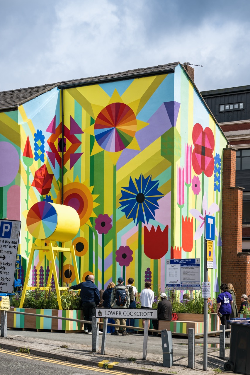 A group of people stands in front of a building with a large, colourful mural of abstract flowers and geometric shapes. The scene includes street signs, a yellow structure, and the street name Lower Cockcroft.
