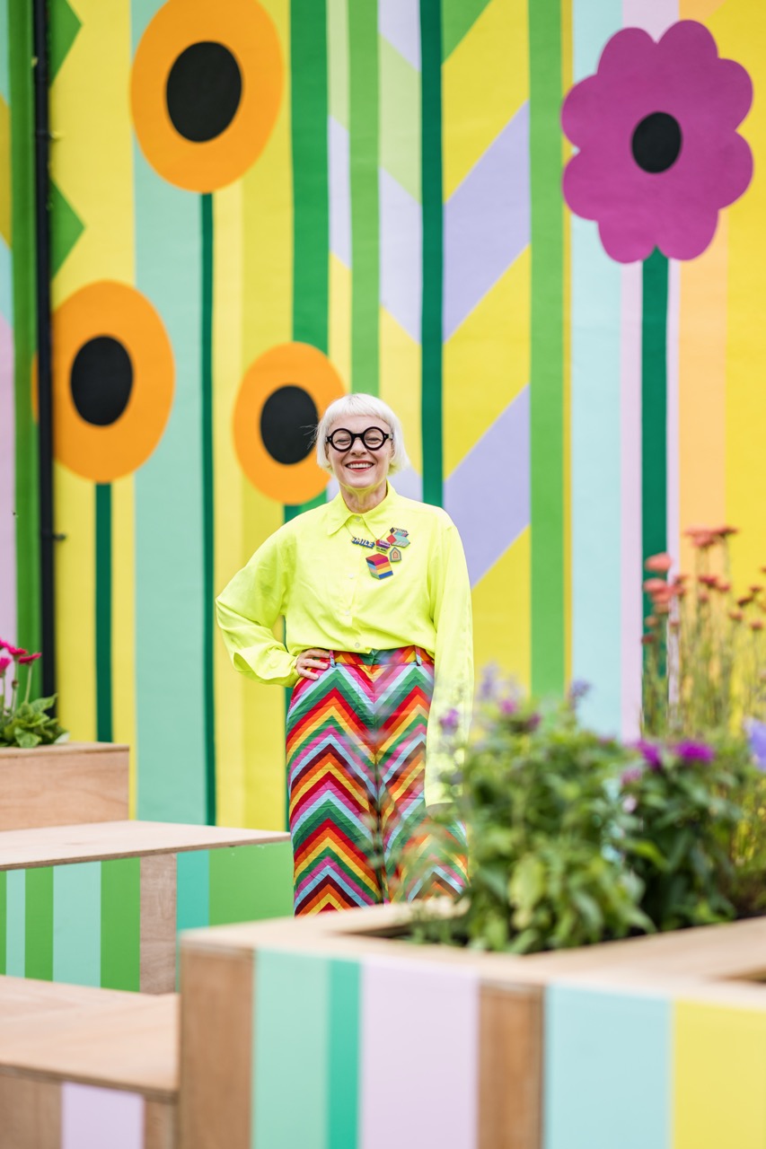 A smiling person with white hair, wearing large glasses, a neon yellow shirt, and colourful chevron trousers, stands in front of a vibrant wall with floral and geometric patterns and surrounded by planters with flowers.