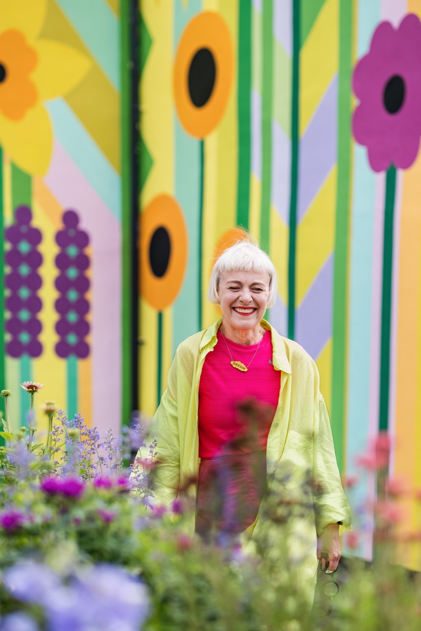A smiling woman with short blonde hair, wearing a bright pink top and a light yellow jacket, stands among colourful flowers in front of a vibrant mural featuring large, stylised flowers and geometric patterns.