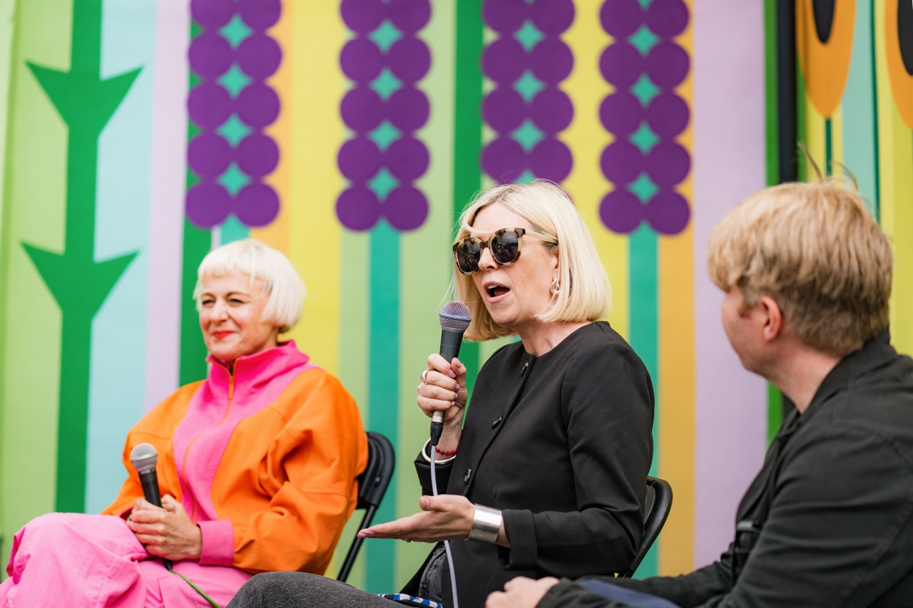 Three people sit on stage with microphones in front of a colourful, patterned backdrop. A woman in orange and pink sits on the left, a woman in black with sunglasses speaks in the centre, and a man in black sits on the right.