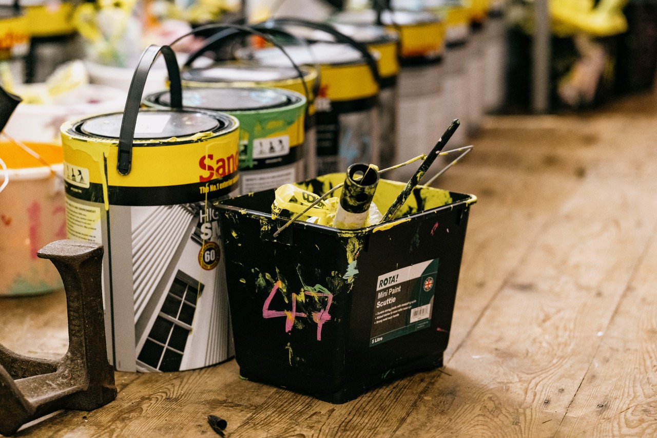 Several paint tins and a black paint scuttle with brushes and a roller, splattered with yellow and green paint, sit on a wooden floor, suggesting an ongoing painting project.
