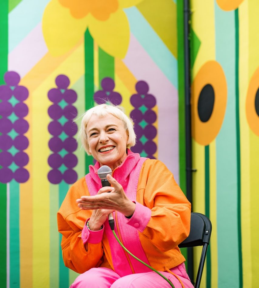 An older woman with short white hair, wearing a bright orange and pink outfit, sits on a chair and smiles whilst holding a microphone. She gestures with one hand in front of a colourful, abstract mural background.