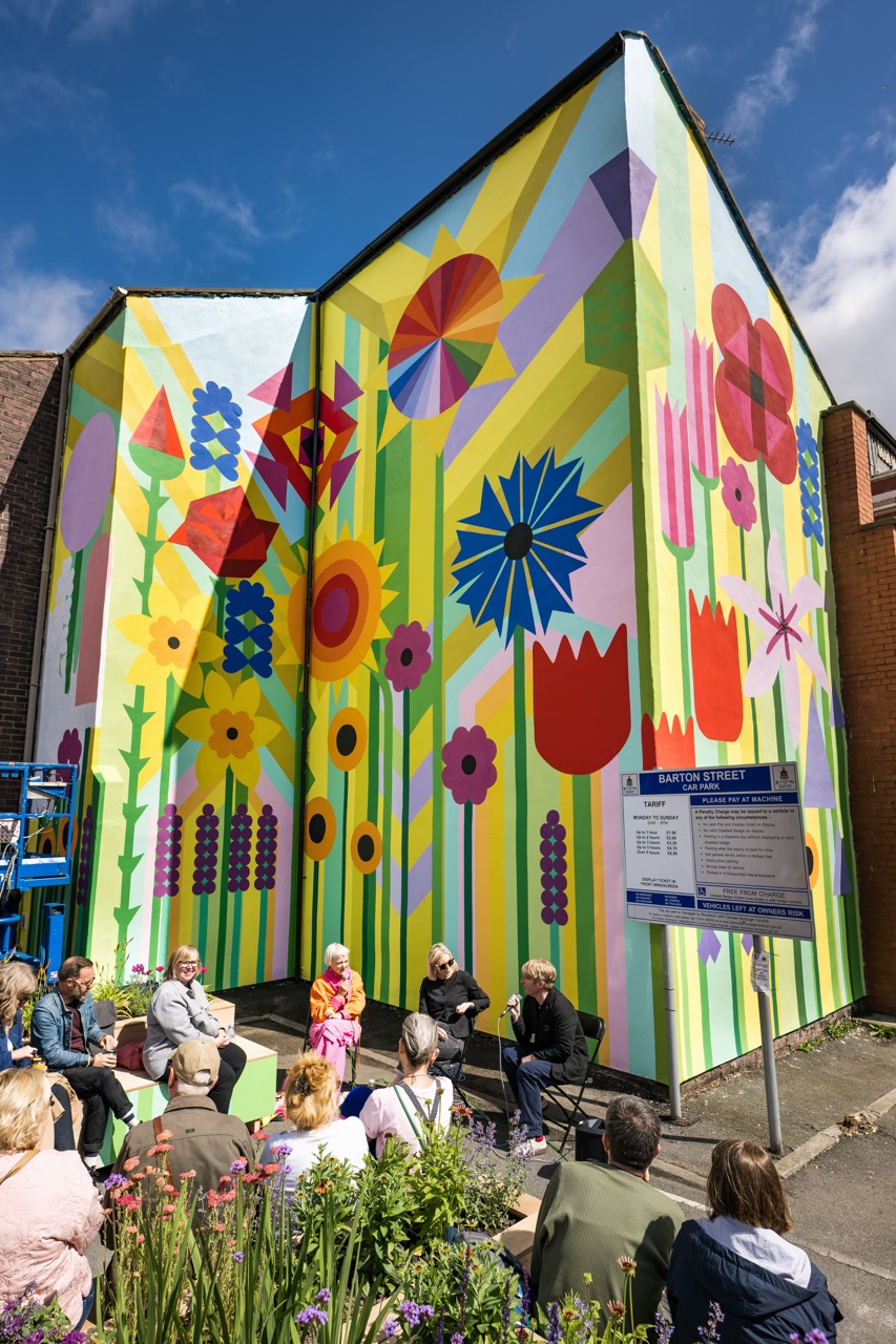 A group of people sits and listens beside a building with a large, colourful mural of abstract flowers and geometric shapes under a bright blue sky. A sign and blooming plants are visible in the foreground.