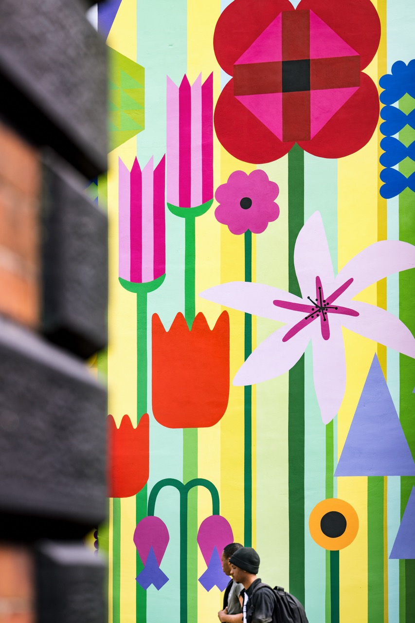 A person walks past a colourful mural featuring large, stylised flowers in red, pink, white, and purple on a yellow and green striped background, partially framed by a blurred brick wall.