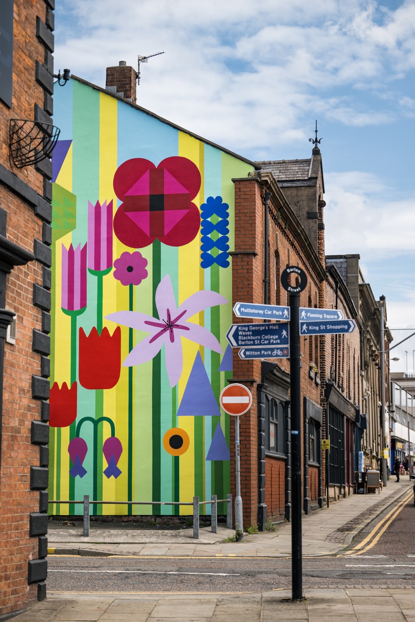 A colourful mural featuring large geometric flowers decorates the side of a building at the end of a street. Road signs and red-brick buildings surround the mural under a partly cloudy blue sky.