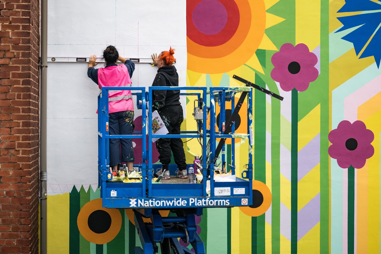 Two people on a blue lift paint a colourful mural with large flowers and geometric shapes on a wall. They work next to a brick building, using tools and wearing casual clothing and safety vests.