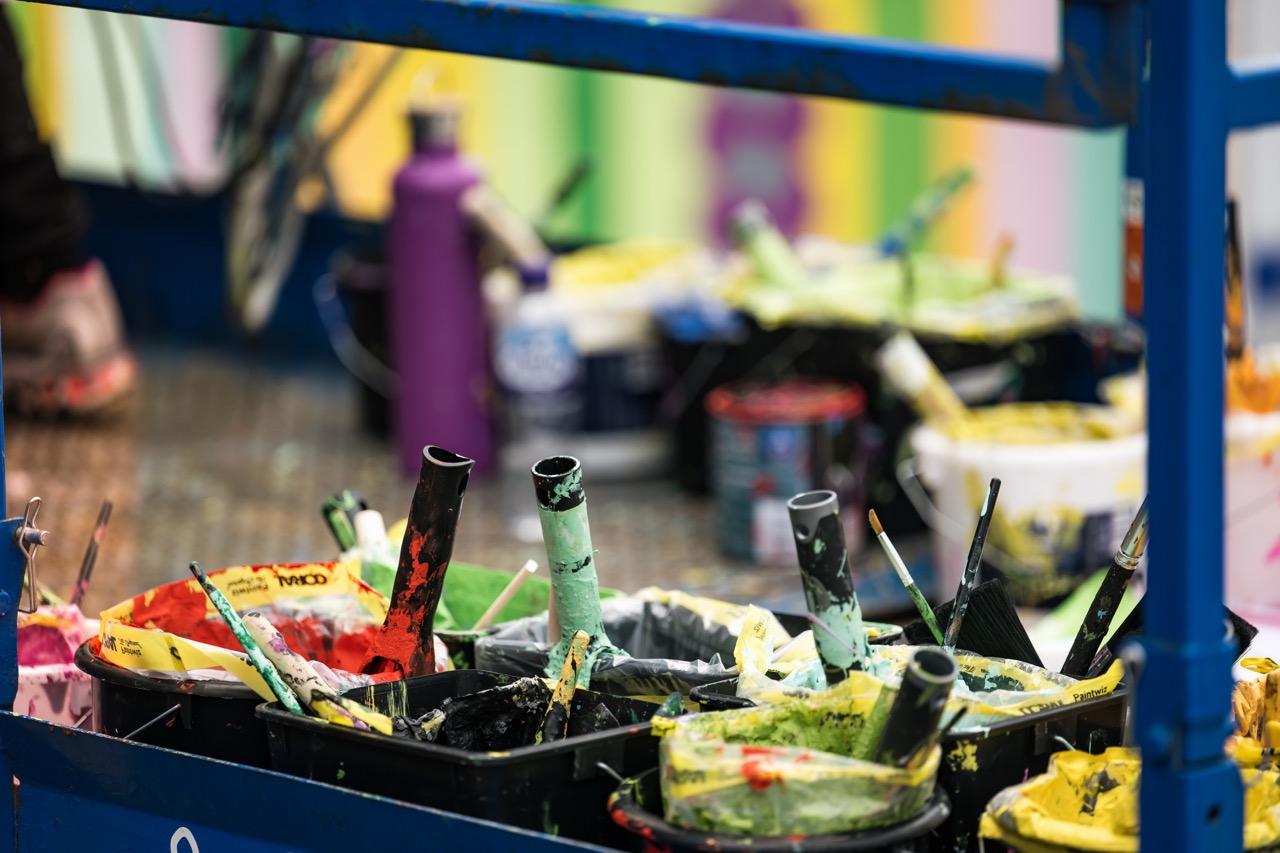 Several paint rollers and brushes covered in colourful paint rest in plastic trays, with paint tins and a purple bottle in the background. The scene suggests an ongoing painting or mural project.