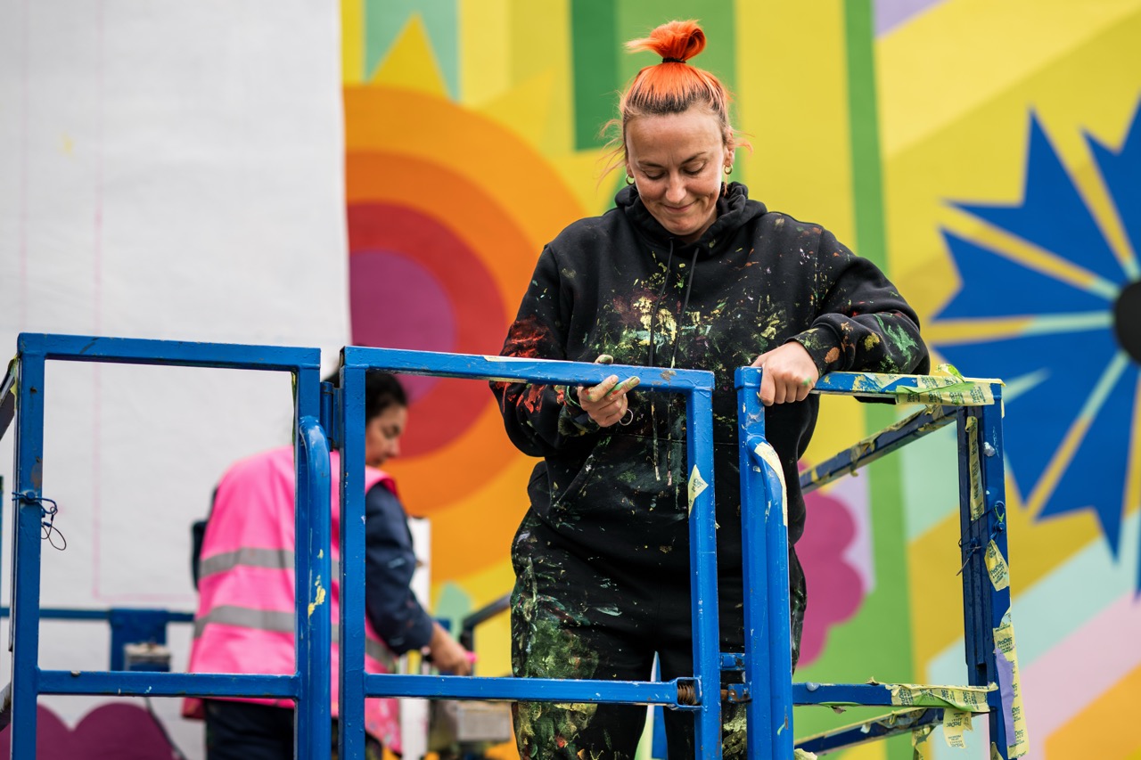 A woman with an orange bun, wearing a paint-splattered black hoodie, stands on a blue lift whilst painting a colourful mural. Another person in a pink vest works in the background. Bright geometric shapes fill the mural.