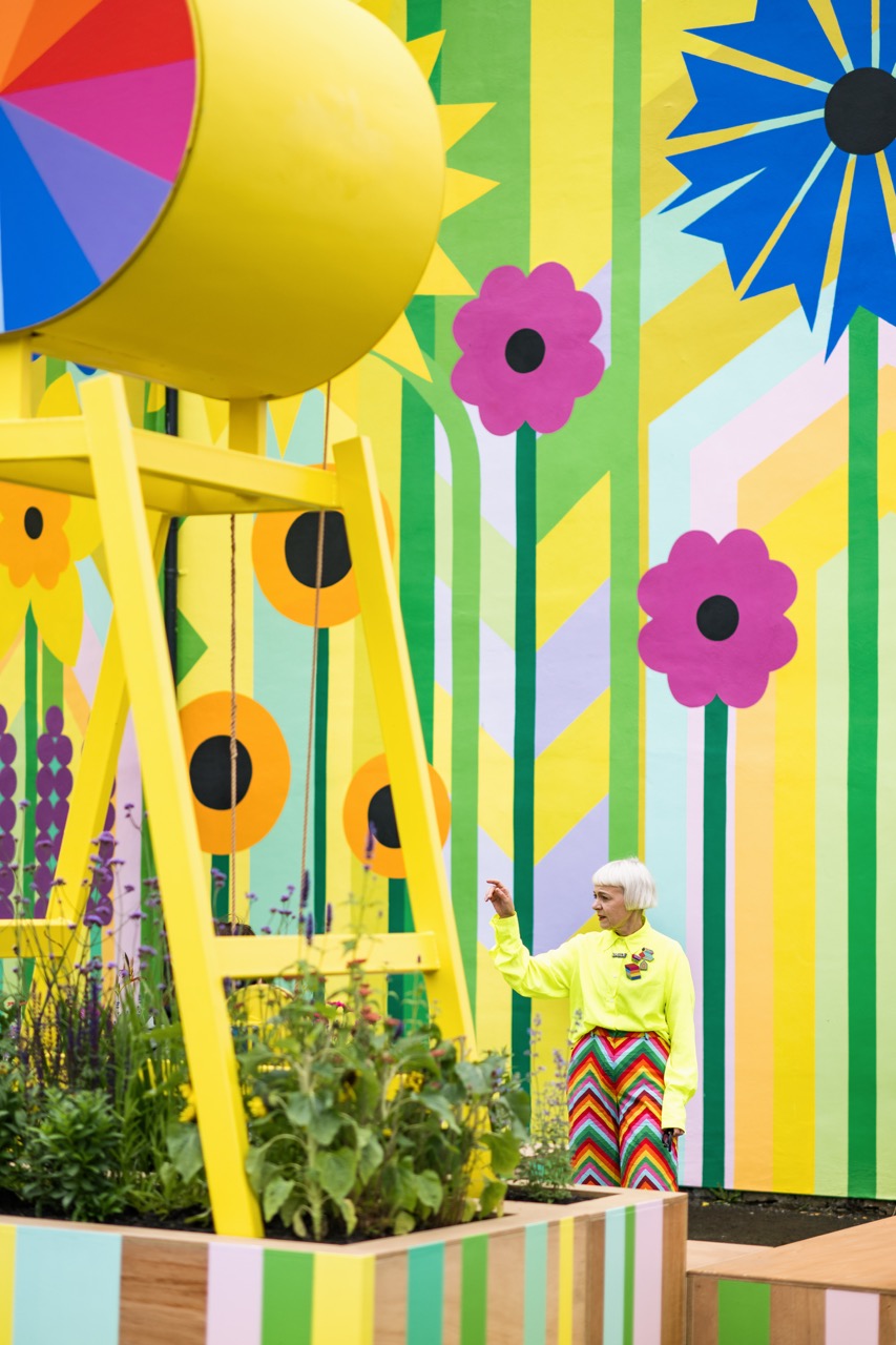 A person in a bright yellow top and colourful zigzag skirt stands near a yellow structure in front of a vibrant mural with large, bold flowers and geometric patterns in yellow, green, pink, and blue tones.