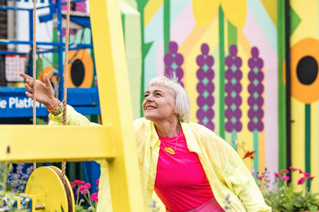 An older woman with short white hair, wearing a bright pink shirt and yellow jacket, smiles and reaches up playfully in front of a colourful mural with floral patterns. Flowers and plants surround her.