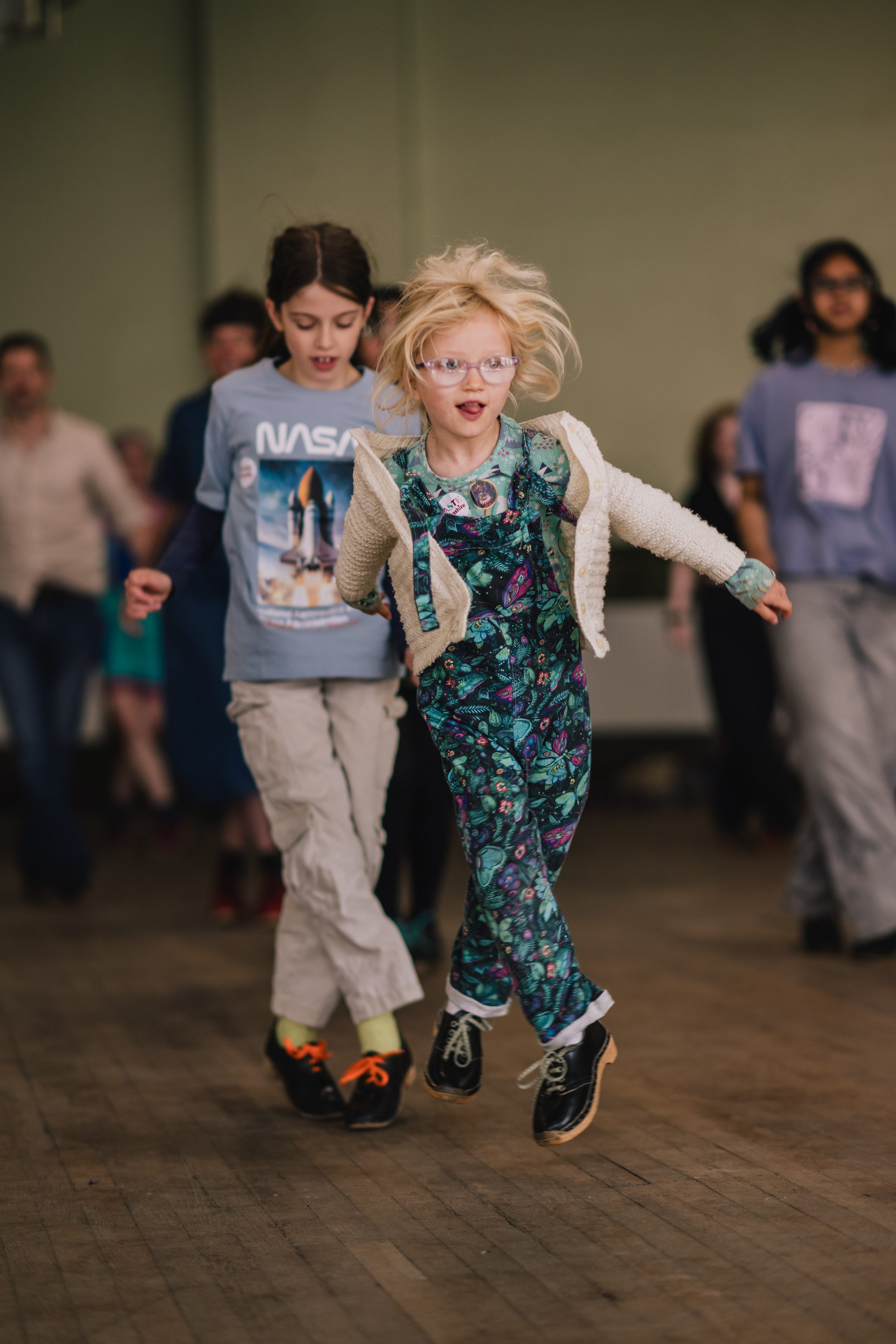 Two children, one in a floral playsuit and glasses, and the other in a NASA t-shirt, joyfully run indoors on a wooden floor, with other people visible in the blurred background.